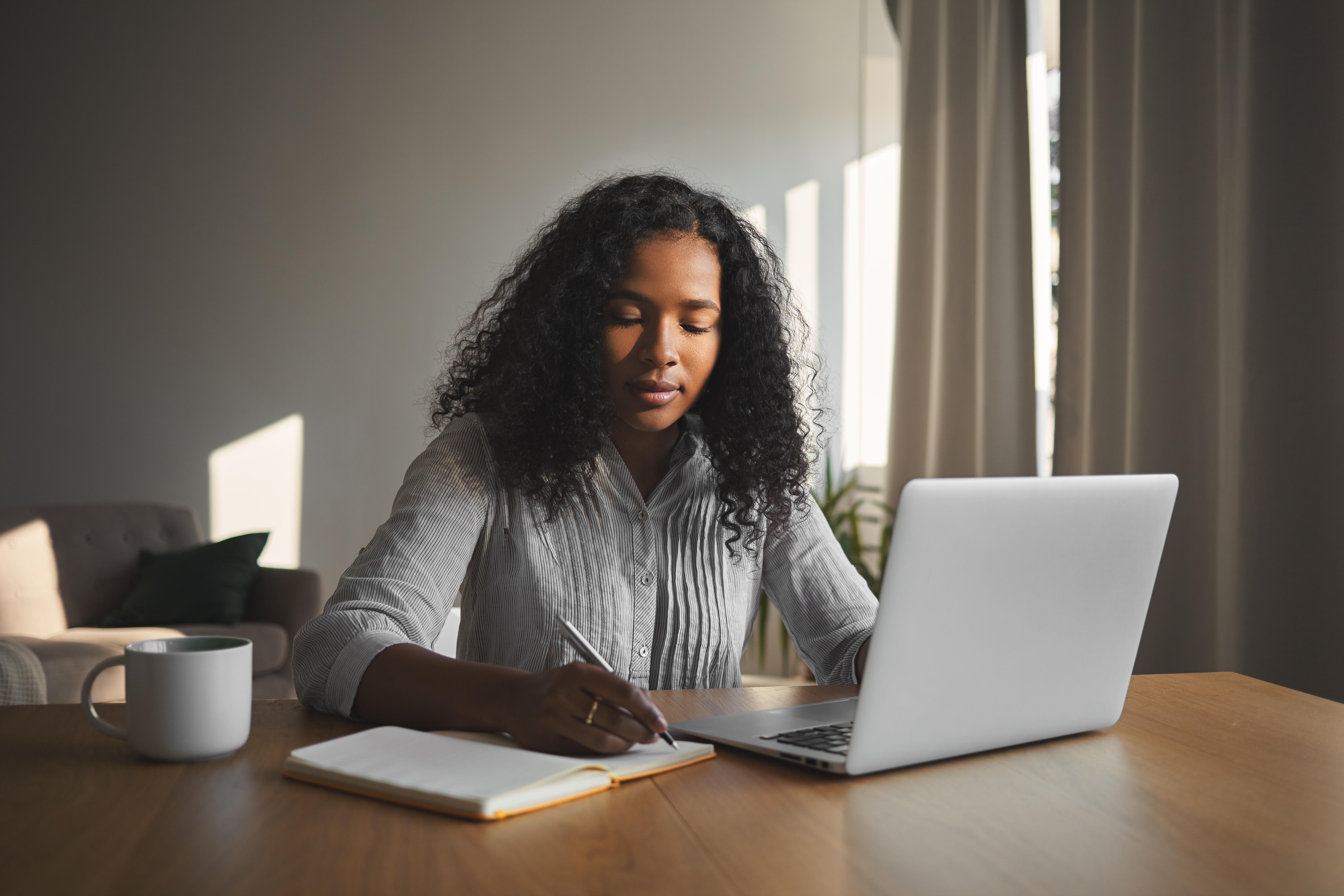 Beautiful mixed race student girl with voluminous hairdo working home assignment her room sitting wooden desk using laptop writing down copybook people technology education